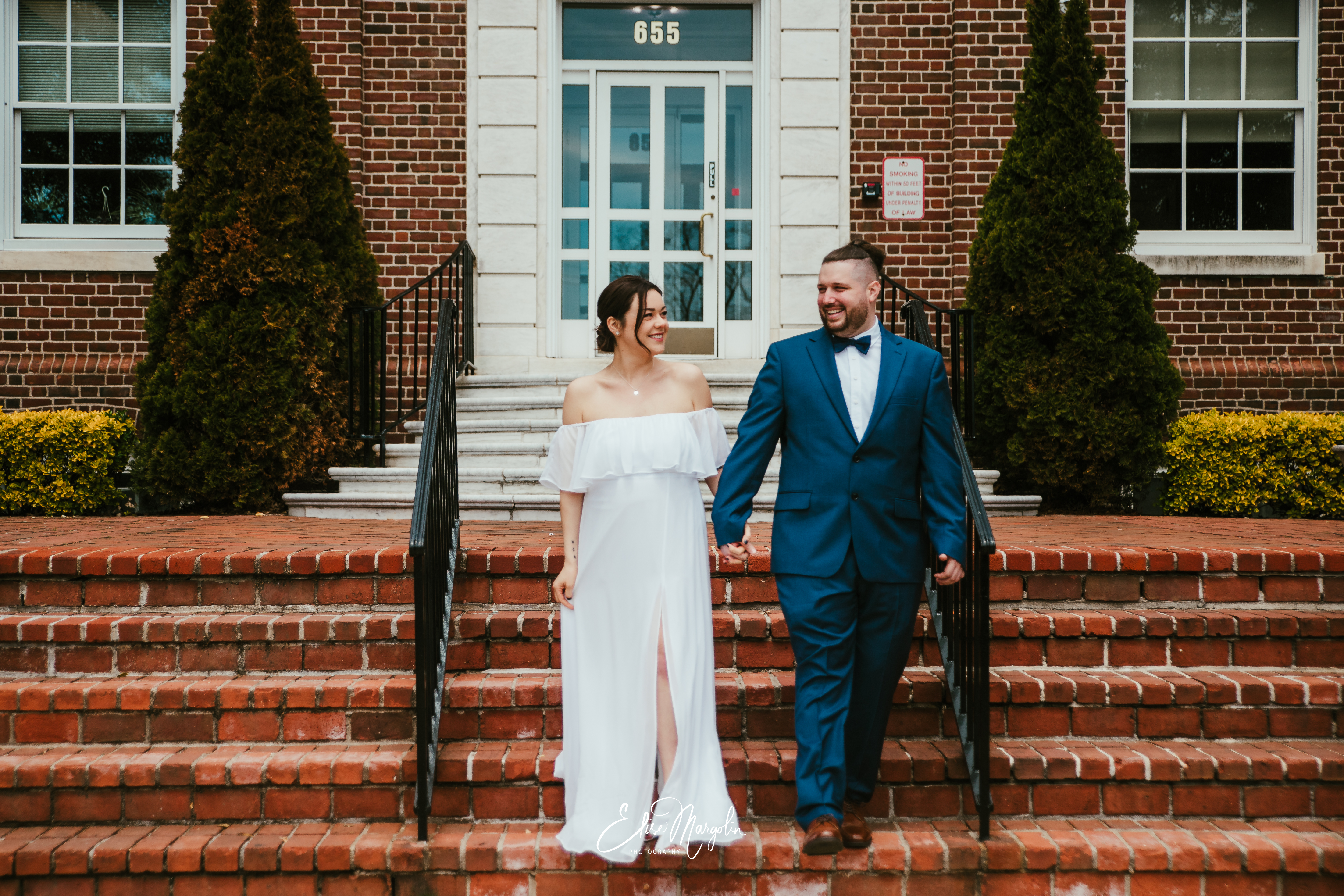 Kristin and Michael holding hands and smiling as they walk down the brick steps outside Islip Town Hall after their intimate wedding ceremony. Kristin is wearing an off-the-shoulder white gown, and Michael is dressed in a navy blue suit with a bow tie.