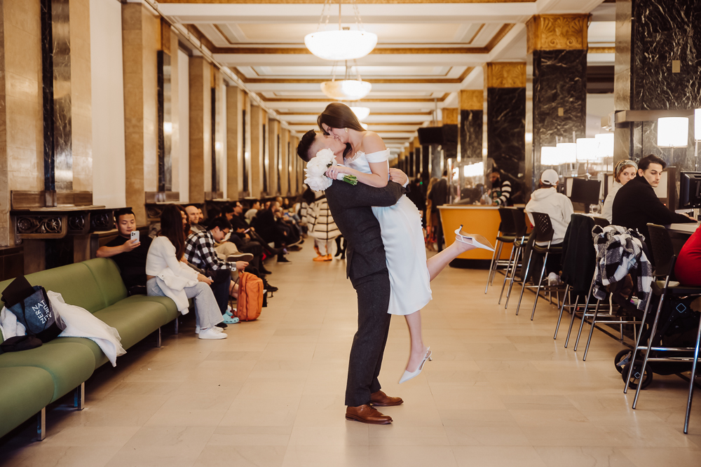 oanna and Taspar sharing a kiss in the hallway of Manhattan City Hall, surrounded by seated guests waiting for their turn. The bride is lifted off the ground by the groom, holding a bouquet of white flowers, capturing a joyous and romantic moment.